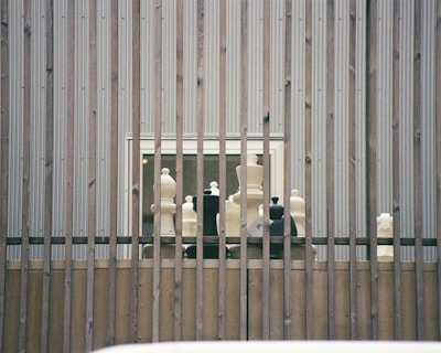 Large chess pieces are arranged on a wooden platform behind vertical metal bars. The setting appears to be outdoors against a corrugated metal wall with two small windows visible.