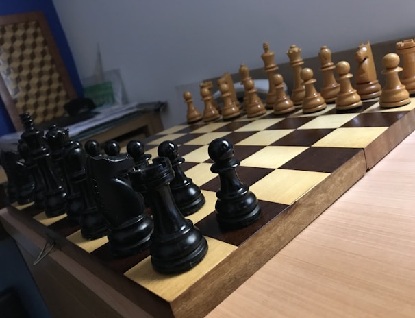 A wooden chessboard is set up for a game with black and white pieces positioned in their starting places. The board is placed on a wooden table, and there is soft lighting in the room. In the background, papers are loosely stacked on a table.