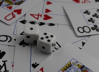 A collection of playing cards spread out, featuring various suits such as hearts, clubs, and spades. Two white dice with black dots are placed prominently on top of the cards.