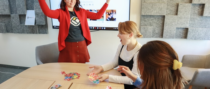 Four people are engaged in a lively game around a wooden table. One person stands with raised arms, while three others are seated, focusing on colorful cards in their hands. There is a computer monitor in the background displaying a webpage, and the room features a modern design with gray acoustic panels on the walls.