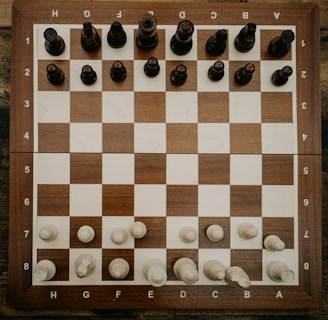 A wooden chessboard is set up with chess pieces in their starting positions. The board is placed on a rustic wooden table, and the chess pieces are made of wood, with darker pieces at the top and lighter pieces at the bottom.