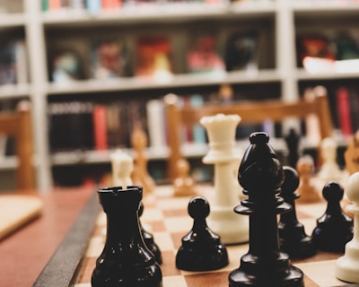 A close-up of a chessboard with black and white chess pieces arranged for play. The background consists of bookshelves filled with various books, creating a cozy, intellectual atmosphere.